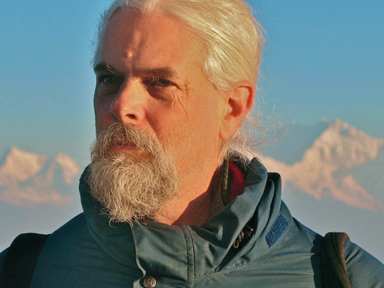 [cropped close-up of man in teal blue windbreaker, with white hair, moustache, and beard, and gazing out pensively, against a background of sunlit snowy mountains and bright blue sky]