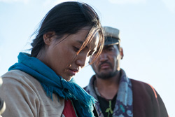 [close up of moustached asian man in brown jacket with blue patterned scarf and white cap with black visor, stands behind and looks sternly at an asian woman with tied dark hair, light tan jacket over red shirt, and blue scarf, while she gazes down forlornly]