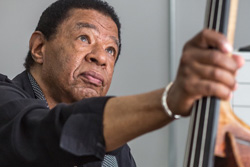 [older black man with light colored eyes wearing a black shirt and black scarf with small white dots gazes upward while holding the neck of a bass with his right hand]