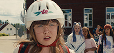 [young white, long blond-haired girl with glasses, and a white bicycle helmet with flower motif at rim, stares out with a puzzled expression, as a group of girls with varied expressions stand to her left amidst a background of dark red building with windows and small white house in distance]
