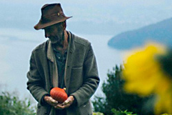 [bearded older man in brimmed brown hat and loose loden-colored jacket over a light tan vest and faded dark blue henley, holds a small dark orange pumpkin in cupped hands, stands gazing down to his right, against a bluish backdrop of lake and hills, and out-of-focus large yellow flower in the right foreground]