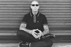 [black and white photo of young man with shaved head and tatooed arms, wearing dark glasses and clothes, sits cross legged with finger tips touching, against a slatted wood background]