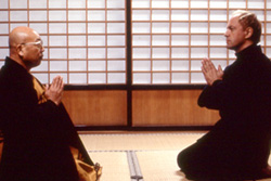 [two men in black kneel on tatami mats facing each other with clasped hands with a shoji screened background]