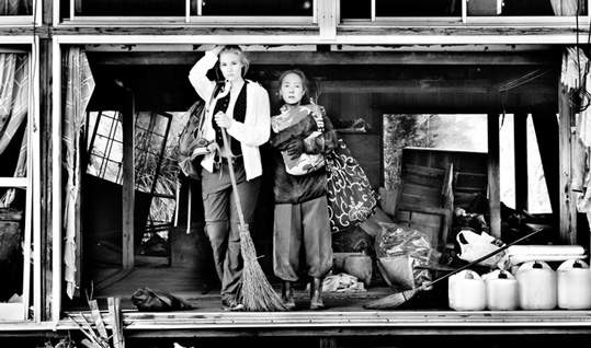 [black and white photo of young blond haired woman holding a broom, with right arm extended overhead, standing next to an older asian woman holding something, standing with a wrecked, open wooden house behind them, surrounded by cleaning supplies]