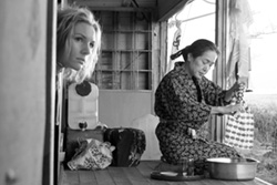 [black and white image of blond young woman gazing out a door alongside an asian woman seated and working outside]