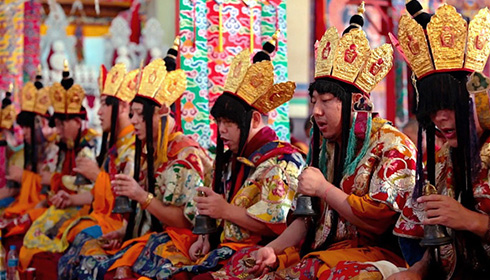 [row of chanting, seated monks with colorful robes and golden crowns with long black hair]