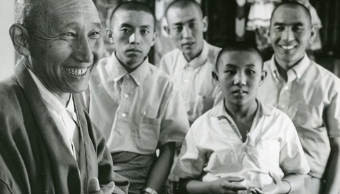 [black and white photo of smiling man with shaved head, wearing a white shirt and grey loose jacket, appearing left of a group of four young men and boys with shaved heads and short sleeved white shirts]