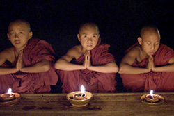 [three young monks in dark red robes sit behind a low wooden table with a burning lamp in front of each of them]