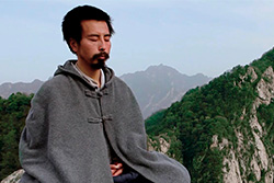 [dark haired man with mustache and goatee, wearing a grey cape, sitting with eyes closed, against a tree capped mountainous background]
