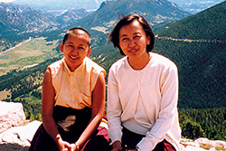 [two smiling women, one with shaved head, sit side by side on a stone wall with mountainous landscape behind them]