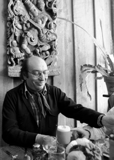 [black and white photo of smiling bald man with glasses sitting at a beverage laden table with wood paneling and Indian art behind him]