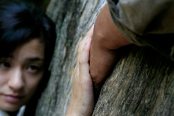 [leaning on a tree trunk, a Japanese woman's face gazes at her hand clasping a man's hand]
