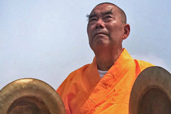 [cropped image of older asian man with shaved head, long eyebrows, and bright orange robe, holding large brass cymbal in each hand]