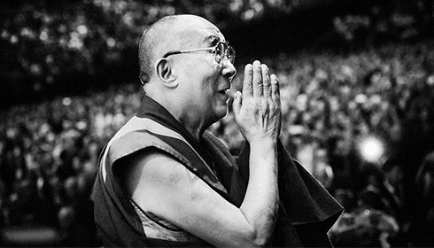 [black and white photo of older man with shaved head, wire rimmed glasses and monk's robe, stands in profile facing right, with raised hands clasped in prayer as he smiles and gazes, with a densely packed, but out of focus background of seated individuals]