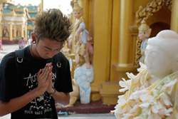 [young asian man with spiked hair wearing a black t-shirt bows head and clasps hands in prayer alongside a white buddha statue draped in white flowers]