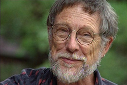[older man with salt and pepper hair, moustache, and beard, wearing thin, circular, wire framed glasses, small gold hoop earrings in his left ear, and a dark gray shirt with light red patterns, smiles to camera, with a lush green background]