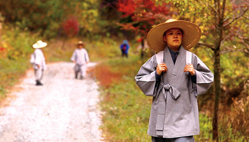 [Asian woman in grey robes and wide brimmed straw hat holds white shoulder straps while walking along a dirt road with autumn colored trees and two smaller figures in background]