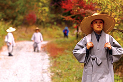 [Asian woman in grey robes and wide brimmed straw hat holds white shoulder straps while walking along a dirt road with autumn colored trees and two smaller figures in background]
