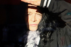 [an older woman with light colored eyes and black hair, wearing a black jacket with white stripes on the cuffs, squints slightly holding up her left arm to block the light in her eyes, with a dark shadow to her right]
