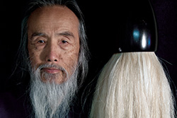 [close-up of white bearded Asian man against a dark background looking to camera with enormous white haired paint brush left of his head]
