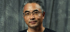 [asian man with salt and pepper hair and eyeglasses with wire frame at top, wearing a black jacket with silver edging, smiling softly to camera, standing against a mottled grey background]