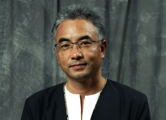 [asian man with salt and pepper hair and eyeglasses with wire frame at top, wearing a black jacket with silver edging against white inset, smiling softly to camera, standing against a mottled grey background]