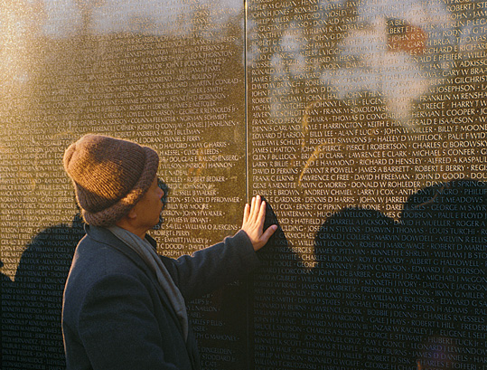 [Thich Nhat Hanh in brown woolen cap and gray coat and scarf places left hand on engraved names on the Vietnam Veterans Memorial Wall with American flag reflected on its surface.]