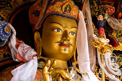 [close up of moustached gold buddha statue head with colored crown and gesturing hand, wrapped in white cloth amidst colorful background]