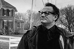 [[black and white image of man in dark framed glasses, dark shirt and jacket looks out frowning with house and grassy area, white fence, and flying flag in background]