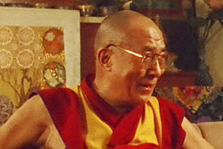 [hh the 14th dalai lama with shaved head, glasses, and red and gold robes, sits against a background of art, smiling while talking]