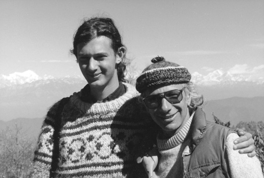 [black and white photo of young man with tied back long dark hair, wearing a thick light colored sweater with dark pattern, with left arm around an older man in woolen hat, light sweater, down vest, and sunglasses, both smiling and gazing to camera with treetops, mountains, and a strip of white clouds in an otherwise clear sky in the background]