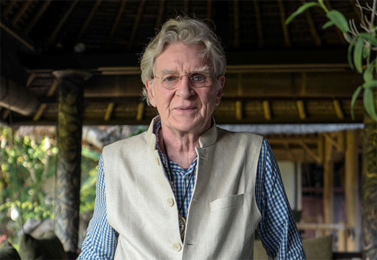 [upper body image of older man with silvery hair, wearing wire rimmed glasses, blue and white checked shirt, and light beige vest, standing in a veranda with brown roof in the background and green tropical leaves in the background]