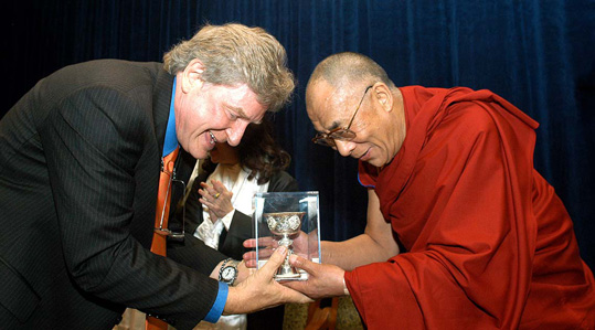 [an older gray haired man at left in dark suit jacket with deep turquoise shirt and light red tie, and an older man in wire rimmed glasses, shaved head, and deep red robe, lean towards each other, smiling, and holding hands supporting a gold and white goblet encased in glass]