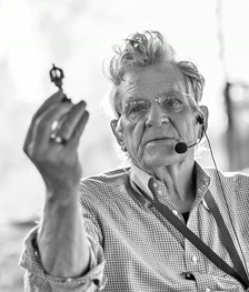 [black and white upper body image of older man with spikey silvery hair, wearing wire rimmed glasses, tiny checked shirt, earset microphone on left ear, and fabric lanyard, holds up a small metal object with his right hand, bent at the elbow]
