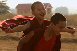 [a laughing young boy with shaved head holds a wind blown maroon fabric in outstretched arms while being carried on the back of a laughing young asian man with dark very short hair and red clothing who is running]