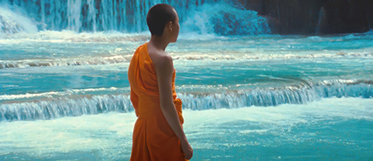 [monk in orange robes with bare right shoulder, and shaved black hair, faces right against a flowing turquoise waterfall background]