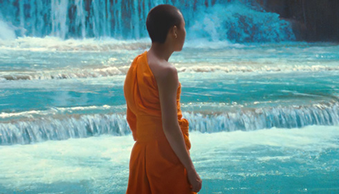 [monk in orange robes with bare right shoulder, and shaved black hair, faces right against a flowing turquoise waterfall background]