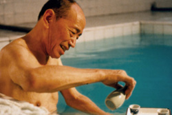 [smiling elderly asian man soaking in white tiled hot tub pours himself a drink]