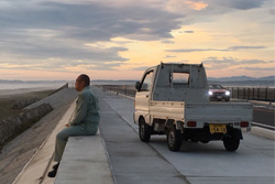 [at dusk, a man dressed in grey, with shaved head and glasses sits at the edge of a road overhang to the left of a small grey truck with a pink lit sky and grey clouds]