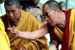 [an asian man with shaved head, metal framed glasses, and red and gold robes, extends his right arm pointing at something while holding a golden metal object in his hands, standing to the left of an asian man draped in darker gold robes and gazing downward]