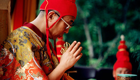 [upper right side of an asian man in gold and red ornately patterned shiny robe and bright red conical fabric hat with straps below his chin, gazes downward towards lifted right hand holding two metal objects held up near his face, against a leafy green background with the head of a man in the same hat blocked by a maroon and gold patterned bowl with blue rim, at camera right]