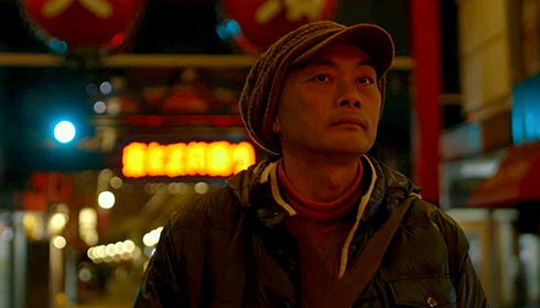 [sad-faced Asian man wearing brown soft cap with visor, pulled down over ears, and layered, matching outerwear, stares out against a nighttime background of brightly lit storefront signs]