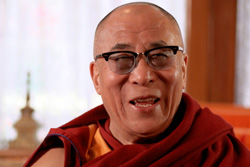 [portrait of a very happy, smiling, older monk, with eye glasses and dark red and gold robe, sitting against a background of a light colored curtained window and tan frame, with small pinnacled statue to the left]