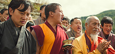 [elderly monk with yellow jacket looks pensive while clapping hands, surrounded to his right by shouting younger monk in gold and maroon robes, holding a small drum-like object with tassels in his left hand, and a male in green jacket looking downward in concern, with other males behind them shouting and clapping]