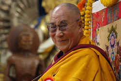 [smiling man with glasses, shaved head, and gold and maroon robes looks to camera while seated near a buddhist statue]