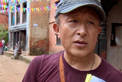 [asian man with blue baseball cap, maroon t-shirt, talks to camera on a street in a dusty town with prayer flags overhead]
