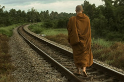 [barefooted man with shaved head, wearing dark orange robes walks with back to camera along railroad tracks]