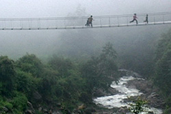 [figure with three children running across a lightweight bridge suspended over a misty verdant landscape with running river]