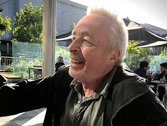 [side profile of smiling older man with grey hair and facial stubble in dark grey shirt and jacket seated in an outdoor eating area with chairs, tables, umbrellas, landscaping, and grey buildings in the background]