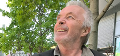[smiling older man with grey hair and facial stubble gazes skyward amidst a background of green leafy trees]